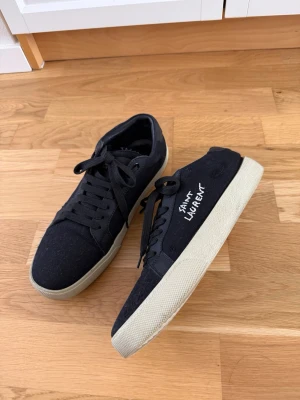 Svarta Y SL sneakers - Säljer ett par svarta sneakers i storlek 41 med vit text på sidan. Skorna har klassisk snörning, svart ovandel i tyg och mocka samt vit sula. Perfekta för dig som gillar stilrena och exklusiva sneakers med en unik touch.