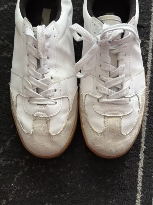 Vita sneakers med mockadetaljer - Snygga vita sneakers med vita skosnören, svart insida och beige mockadetaljer på tån. Yttermaterialet är en mix av läder och mocka, och sulan är i gummi. Perfekta för en clean och stilren look.