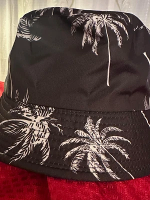 Ny Svart bucket hat med palmtryck  - Snygg svart bucket hat med vita palmträd som tryck runt om. Hatten är tillverkad i 100% polyester och har en avslappnad passform. Perfekt för att addera lite sommarvibbar till din outfit.