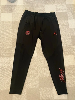 Svarta PSG x Jordan mjukisbyxor - Snygga svarta mjukisbyxor från Jordan i samarbete med PSG. Byxorna har röda detaljer, PSG-logga och Jumpman-logga framtill samt texten 'Paris' på benet. Tillverkade i mjukt material med dragsko i midjan och smal passform.