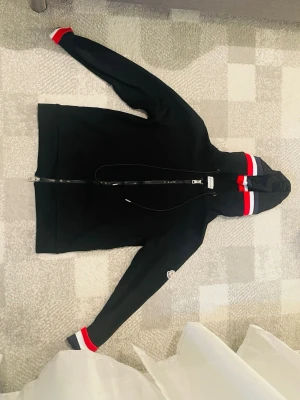 Svart Moncler hoodie med dragkedja - Svart hoodie från Moncler med hel dragkedja och huva. Röd och vit rand på huva och ärmslut ger en sportig vibe. Moncler-logga på vänster ärm. Perfekt för dig som gillar stilrena och exklusiva plagg.