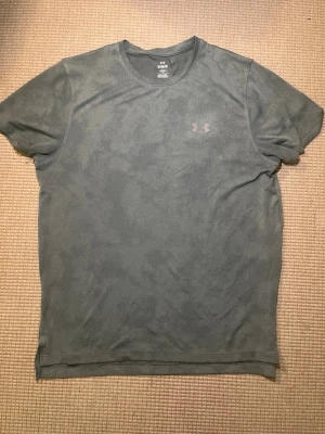 Grå kortärmad t-shirt från Under Armour - Snygg grå t-shirt från Under Armour med diskret logga på bröstet. Klassisk rund halsringning och korta ärmar. Tillverkad i ett lätt och funktionellt material som passar perfekt till träning eller vardagsstil.