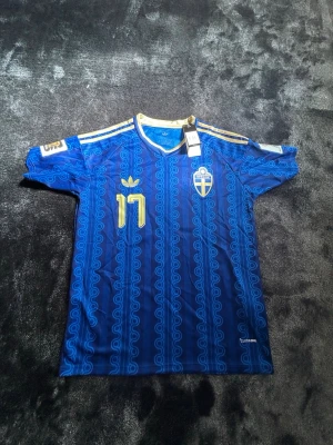 Blå Sverige matchtröja Adidas - Säljer en blå matchtröja från Adidas med Sveriges landslagsemblem och nummer 17 i gult. Tröjan har guldfärgade detaljer, klassiska Adidas-ränder på axlarna och ett unikt mönster över hela tyget. Perfekt för fotbollsfans!