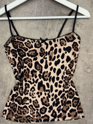 Leopardmönstrad linnetopp från Zara - Säljer en snygg leopardmönstrad linnetopp från Zara med tunna axelband och liten rosett framtill. Toppen har svarta spetsdetaljer längs kanten och är i ett mjukt, stretchigt material. Perfekt för dig som vill sticka ut med ett djurmönster!