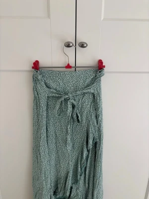 Grön omlottkjol med knyt och mönster - Lång grön omlottkjol med vitt blomster mönster och knytband i midjan. Kjolen har en luftig och flowy passform, perfekt för varma dagar. Materialet känns lätt och bekvämt mot huden. Kjolen är köpt utomlands och har ingen etikett. Det står därför ingen storlek men jag uppskattar att den passar xs/s.