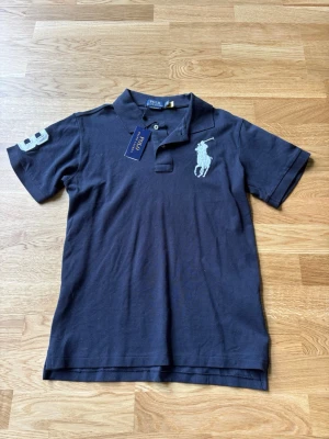 Marinblå pikétröja från Polo Ralph Lauren - Klassisk marinblå pikétröja från Polo Ralph Lauren med stor vit broderad logga på bröstet och siffran 3 på ärmen. Tröjan har krage, knappar vid halsen och är gjord i bomull. Perfekt för en stilren och sportig look.