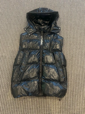 Svart dunväst med huva från TPDY - Snygg svart dunväst från TPDY med glansig finish och stor huva. Västen har dragkedja framtill, två sidofickor och en modern, puffig look. Perfekt för lager på lager och streetstyle. Märkesdetalj på bröstet för extra vibe.