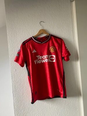 Manchester United matchtröja röd - Säljer en röd Manchester United matchtröja från Adidas med klubbmärke på bröstet och TeamViewer-logga framtill. Tröjan har svarta detaljer på axlar och sidor, samt Premier League-märke på ärmen. Tillverkad i lätt och ventilerande material.