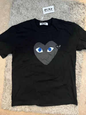 Svart Comme des Garçons Play t-shirt - Säljer en svart t-shirt från Comme des Garçons Play med stort hjärta och blå ögon på bröstet samt ett litet svart hjärta bredvid. Klassisk rund hals och korta ärmar. Perfekt för dig som gillar streetwear och vill sticka ut.