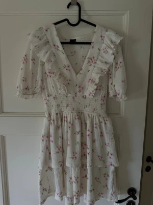 Vit blommig kortklänning med volang - Supersöt vit kortklänning med rosa blommor, volangdetaljer och v-ringning. Klänningen har puffiga korta ärmar och smock i midjan för en bekväm passform. Perfekt för sommarens alla tillfällen!