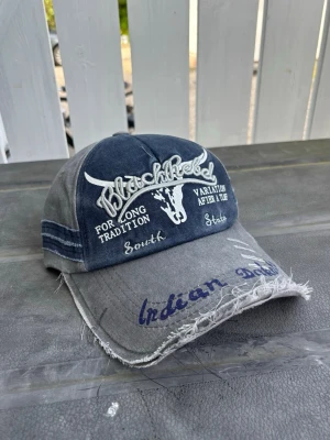 Black rebel vintage distressed retro Grå och blå keps med brodyr - Säljer en grå och blå keps med broderad text och motiv framtill. Kepsen har slitna detaljer på skärmen och texten 'Black Rebel', 'Indian Dakota' samt ett tjurhuvud broderat. Justerbar baktill och tillverkad i bomullstyg.