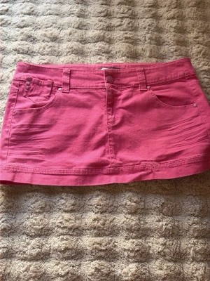 Rosa kort jeanskjol med fickor - Snygg rosa kortkjol i jeansstil med fem fickor och bälteshällor. Kjolen har klassisk knapp- och dragkedjestängning framtill och dekorativa sömmar på bakfickorna. Perfekt för dig som vill ha en färgstark och trendig look.