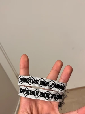 Christian Dior J'Adior tygarmband - Säljer ett snyggt tygarmband från Christian Dior med texten 'J'Adior' i svart på vit bakgrund. Armbandet har en vävd design med grafiskt mönster och fransar i ändarna. Perfekt accessoar för att lyfta din outfit.