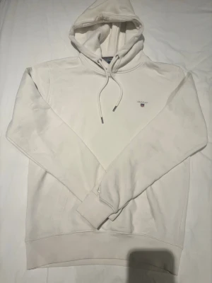 Vit hoodie från GANT med broderad logga - Säljer en stilren vit hoodie från GANT med broderad logga och sköld på bröstet. Hoodien har dragsko i huvan, ribbade muddar och är tillverkad i mjukt bomullsmaterial. Perfekt för en clean och avslappnad look.