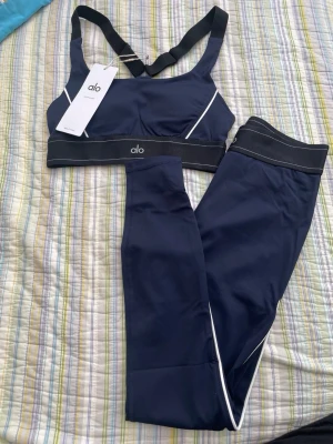 Mörkblått träningsset från Alo - Snyggt mörkblått träningsset från Alo med sport-bh och matchande tights. Setet har vita detaljer och breda svarta resårband med Alo-logga. Materialet är stretchigt och passar perfekt för gym eller yoga.