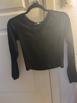 Svart croppad topp från Bershka - Snygg svart croppad topp från Bershka med lång ärm och v-ringad hals. Perfekt till jeans eller kjol för en clean och enkel look. Tillverkad i mjukt material som sitter skönt på kroppen.