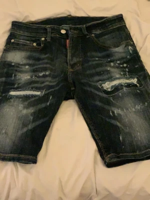 Mörkblå distressed jeansshorts Dsquared2 - Säljer ett par mörkblå jeansshorts från Dsquared2 med slitningar och distressed detaljer framtill. Shortsen har klassisk femficksdesign, knappgylf och vita färgstänk för en edgy look. Perfekta för dig som gillar streetwear och vill sticka ut.