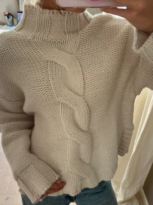 Beige stickad tröja med flätmönster - Mysig beige stickad tröja med hög krage och ett stort flätmönster framtill. Tröjan har breda ribbade muddar vid ärmslut och nederkant samt en avslappnad passform. Perfekt för kalla dagar och enkel att styla med jeans.