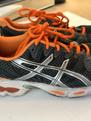 Svarta och orange Asics sneakers - Snygga sneakers från Asics i svart mesh med orange detaljer och snören. Skorna har en vit och orange sula samt den klassiska Asics-loggan i silver på sidan. Perfekta för dig som gillar sportig stil och vill sticka ut lite extra.