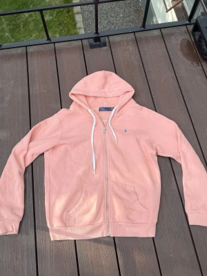Ljusrosa hoodie från Polo Ralph Lauren - Säljer en ljusrosa hoodie från Polo Ralph Lauren med dragkedja, vit dragsko och två fickor framtill. Klassisk blå logga broderad på bröstet. Tillverkad i mjukt bomullsmaterial och har ribbade muddar vid ärmslut och nederkant.
