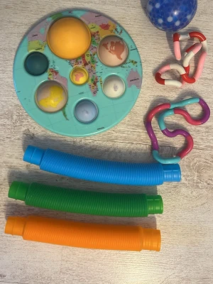 Fidgetleksaker mixad samling - En samling fidgetleksaker med blandade färger och former, inklusive en pop it med världskarta, tre pop tubes i blått, grönt och orange, en blå stressboll med vita prickar samt två olika tangle-leksaker i rosa, rött, vitt, lila och turkost. Perfekt för att hålla händerna sysselsatta.