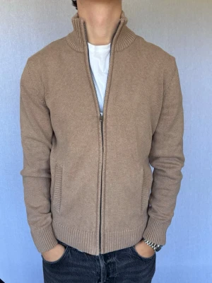 Beige stickad ziptröja från Dressmann  - Detta är en kofta från Dressmann i storlek S med normal passform. Modell på bild är ca 180, 66kg. Plagget är i bra skick utan några defekter. Hör gärna av dig vid intresse eller frågor! 