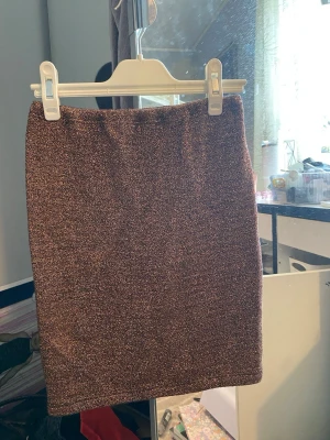 Glittrig brunguldig kortkjol - Säljer en glittrig brun kortkjol med rak passform. Kjolen har en elastisk midja och materialet skimrar snyggt i ljuset. Perfekt för dig som vill sticka ut lite extra med en enkel men ändå festlig look.