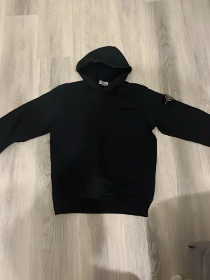 Svart hoodie från Stone Island - Säljer en svart hoodie från Stone Island med den klassiska loggan på vänster ärm. Hoodien har en enkel och stilren design med huva och ribbade muddar. Perfekt för dig som gillar streetwear och vill ha något bekvämt och snyggt.