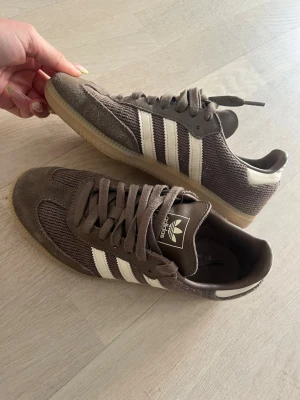 Adidas Samba Og - Snygga bruna Adidas samba og i färgen earth strata med klassiska vita ränder på sidorna och gummisula. Ovandelen är i mocka och mesh för en sportig och retro vibe. Perfekta för dig som gillar streetstyle och vill ha något bekvämt och stilrent till vardags. Köpa från Nelly för 1499