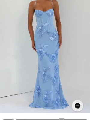 Ljusblå lång klänning med blommor - Supersnygg ljusblå lång klänning med tunna axelband och figurnära passform. Klänningen har vackra 3D-blommor i spets över hela tyget och en markerad byst med corsettdetaljer. Perfekt för bal eller speciella tillfällen.