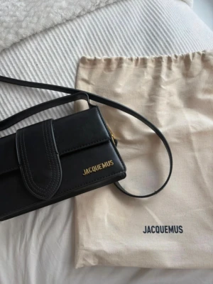 Svart Jacquemus handväska i läder - Snygg svart handväska från Jacquemus le bambino den stora i slät läderlook med guldiga detaljer och logga framtill. Väskan har både axelrem och handtag, samt en praktisk ficka på baksidan. Perfekt för dig som gillar stilrena accessoarer med lyxig känsla.