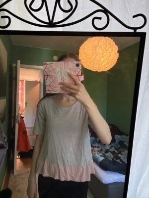Grå topp med rosa volangkant - Söt och avslappnad grå topp med ljusrosa volangkant nertill. T-shirten har rund halsringning och korta ärmar. Perfekt för en chill och bekväm stil. I 98% bomull och 2% viskos 