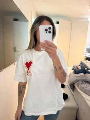 Vit t-shirt från Ami Paris med hjärtdetalj - Säljer en vit t-shirt från Ami Paris med en stor röd hjärtdetalj och bokstaven A på bröstet. T-shirten har normal passform, rund hals och är kortärmad. Tillverkad i mjuk bomull som känns skön mot huden. Perfekt för en clean och trendig look. Står ingen storlek men skulle säga är S/M och de är unisex 