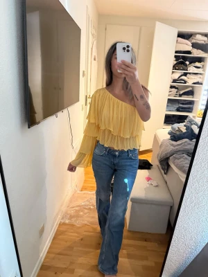 Gul volangig oneshoulder topp - Snygg gul topp med volanger och plisserat tyg. Toppen är oneshoulder med lång ärm på ena sidan och ärmlös på andra. Croppad modell som ger en trendig look, perfekt för dig som vill sticka ut.