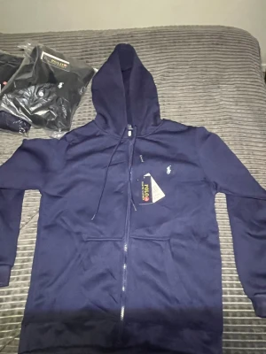 Mörkblå hoodie från Polo Ralph Lauren - Snygg mörkblå hoodie med dragkedja från Polo Ralph Lauren. Klassisk design med vit broderad logga på bröstet, huva med dragsko och två fickor framtill. Perfekt för en avslappnad och stilren look. Passar S och M