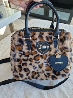 Juicy Couture leopardmönstrad handväska - Superfin handväska från Juicy Couture i fluffigt fuskpälsmaterial med leopardmönster. Väskan har svarta handtag och axelrem i läderimitation samt hjärtformat emblem och tag med Juicy Couture-logga och krona.