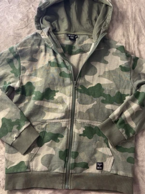 Klassisk camo hoodie med dragkedja - Säljer en grön camouflagemönstrad hoodie från Lager 157 med dragkedja framtill och två fickor. Hoodien har ribbade muddar vid ärmslut och nederkant samt en skön huva. Perfekt för dig som gillar streetwear och vill ha en avslappnad stil.