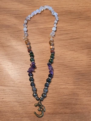 Färgglatt halsband med Om-symbol - Unikt halsband med halvädelstenar, inklusive genomskinliga, gröna, lila och bruna stenar. Halsbandet har en metallberlock i form av aum-symbolen. Perfekt för dig som gillar boho-stil och vill ha något personligt och färgstarkt. Halsbandet kommer i en söt liten ask tillsammans med information om kristallernas magiska egenskaper. 