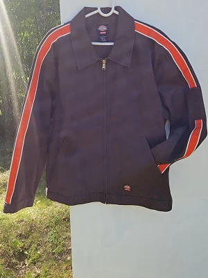 Dickies x Supreme  jacka - Svart jacka från Dickies x Supreme med röda och vita ränder längs ärmarna. Jackan har dragkedja framtill, två snedställda fickor och klassisk krage. Materialet är slitstarkt och perfekt för streetwear-stil.
