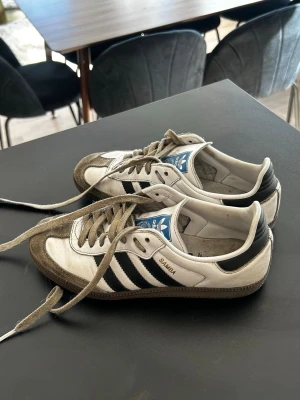 Adidas Samba vita och svarta sneakers - Klassiska Adidas Samba sneakers i vitt och svart med bruna detaljer. Skorna har den ikoniska tre ränder-designen på sidan, blå logga på plösen och texten 'SAMBA' i guld. Ovandelen är i läder och mocka, med gummisula och lågt skaft.
