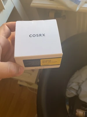 COSRX Advanced Snail 92 All in one Cream - En vit förpackning med COSRX Advanced Snail 92 All in one Cream, en populär ansiktskräm med snigelsekret. Krämen är känd för att återfukta och stärka huden. Förpackningen innehåller 100 g och har en stilren design med svart och gul etikett.