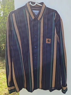Randig skjorta från Carhartt - Snygg långärmad skjorta från Carhartt med vertikala ränder i marinblått, beige och grönt. Skjortan har klassisk krage, knäppning framtill och en bröstficka med Carhartt-logga. Perfekt för en stilren och avslappnad look.