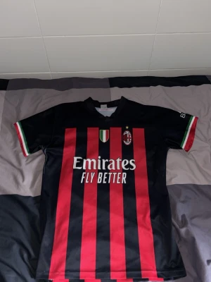 AC Milan matchtröja Ibrahimović 11 - Snygg AC Milan matchtröja med röda och svarta ränder, klubbmärke och sponsortryck på framsidan. På ryggen står Ibrahimović och nummer 11. Tröjan har italienska flaggans färger på ärmsluten och är tillverkad i lätt och ventilerande material.