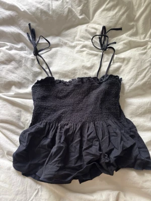 Svart linnetopp med knytband - Snygg svart linnetopp med smockad överdel och volang nertill. Axelbanden är tunna och knyts upptill för en justerbar passform. Perfekt till jeans eller kjol för en trendig look.