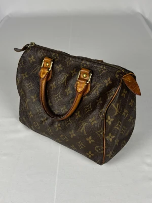 Louis Vuitton Speedy 25 - Louis Vuitton Speedy handväska med det klassiska monogrammet. Size 25 (den är 25 cm bred). Väldigt fint skick överlag men har två mindre defekter. Dragtappen sitter löst och rammlar av om man drar hårt i den (bild 6), den fungerar fortfarande att använda och om man vill så går det bra att använda väskan utan dragtappen. Det finns även ett väldigt litet hål i lädret (bild 7), ungefär en halv millimeter stort och omöjligt att notera vid använding. Utöver det väldigt välbehållen och fint skick. 
