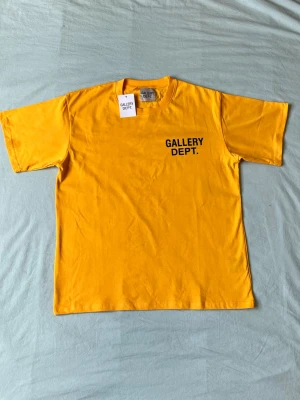 Gul t-shirt från Gallery Dept. - Gul t-shirt från Gallery Dept. med svart tryck på bröstet. Klassisk rund halsringning och korta ärmar. Tillverkad i mjuk bomull för en skön och avslappnad stil.