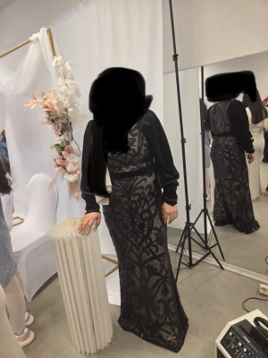 Svart långklänning med mönster - Elegant svart långklänning med transparenta partier och dekorativt sammetsmönster. Klänningen har lång ärm och markerad midja, vilket ger en snygg siluett. Perfekt för dig som vill sticka ut med en unik och stilren look. Använd bara 1 gång