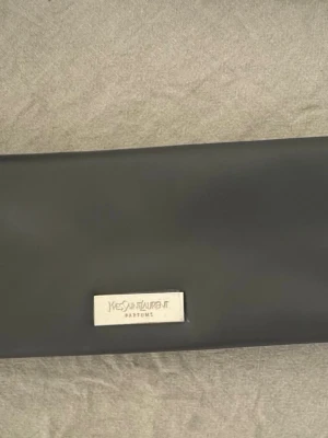 Ysl grå kuvertväska  - Stilren grå kuvertväska från Yves Saint Laurent med en rektangulär form och en silverfärgad metallbricka framtill med märkets logga. Väskan är tillverkad i ett slätt material som ger en lyxig känsla.
