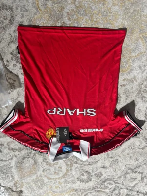 Manchester United röd t-shirt Umbro - Klassisk röd Manchester United t-shirt från Umbro med vit krage och svarta samt vita detaljer på ärmarna. Tryck med SHARP och klubbmärke framtill. Perfekt för fotbollsfans som vill sticka ut med retrostil.