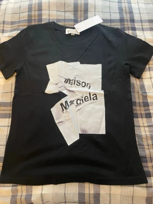 Maison Margiela svart t-shirt - Svart t-shirt från Maison Margiela med ett unikt grafiskt tryck i vitt och svart framtill. T-shirten har korta ärmar. Tillverkad i mjuk bomull för en skön känsla och modern look.